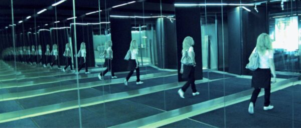 Blonde Frau in einem Durchgang, auf beiden Seiten Spiegel, deswegen "endless mirror"-Effekt.